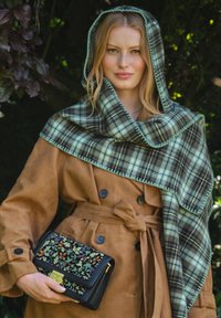 Manteau trench en suede marron avec une taille nouée, associé à un châle à carreaux vert et noir. Pochette noire ornée d'une broderie florale colorée.