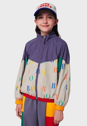 Veste coupe-vent colorée en violet, crème et vert, ornée de lettres aléatoires dans différentes couleurs, avec un ourlet élastiqué et des poignets élastiques.