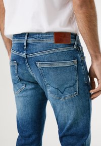 Denim jeans in medium blue with a slight fade, featuring a brown leather brand patch, back pockets with stitching detail, and a straight cut.