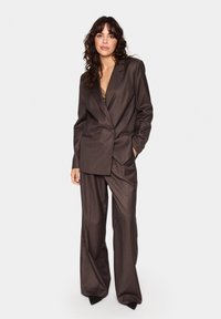 Costume marron sur mesure avec un blazer croisé à revers et un pantalon à jambes larges. Texture lisse et design sophistiqué.