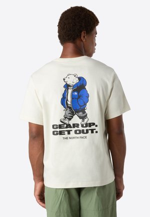 Mand med grønne shorts iført hvid T-shirt med grafik af en gående bjørn i blå jakke og teksten "Gear Up. Get Out. The North Face" på ryggen.