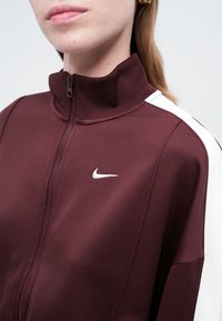 Burgundská zipsová bunda s vysokým golierom, s bielymi pruhmi na pleciach a malým bielym logom Nike na hrudi. Hladká textúra látky.