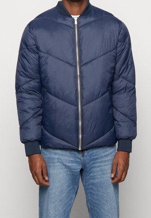 Marineblauer Pufferjacke mit Zickzack-Steppung, einem Reißverschluss vorn, gerippten Bündchen und Kragen, aus einem glatten, glänzenden Stoff. Über Jeans getragen.