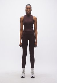 Γυναίκα με σκούρο ρούχο γυμναστικής που κρατά μικρά αλτήρες, στέκεται όρθια μπροστά σε λευκό φόντο.