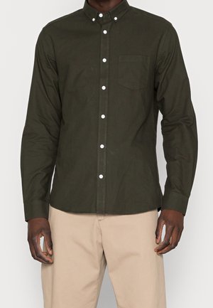 Homme portant une chemise à manches longues verte foncée boutonnée avec une poche poitrine et un pantalon beige, debout devant un fond uni.