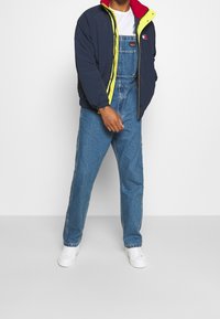 Gewatteerd jack in marineblauw met rode en gele accenten, gecombineerd met blauwe denim tuinbroek. De outfit wordt aangevuld met witte sneakers.