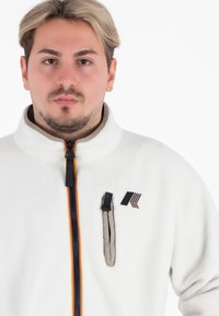 Biała polarowa kurtka z wysokim kołnierzem, przednim zamkiem błyskawicznym, kontrastującym zamkiem w kolorze granatowym i pomarańczowym oraz kieszenią na lewej piersi z logo i dodatkowymi akcentami.