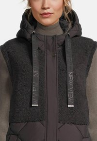 Femme portant un gilet noir matelassé avec des panneaux en polaire et une capuche, par-dessus un pull à col roulé gris côtelé.