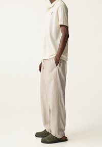 Hellbeige Kurzarm-Polohemd aus strukturiertem Stoff mit einem orangefarbenen Ananas-Graphic, kombiniert mit lockeren hellgrauen Hosen und olivgrünen Clogs.