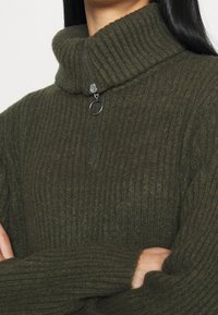 Olivengrønt ribbet rullekrave-sweater med halv lynlås og en sølv lynlås. Blød tekstur, løst fit og let overdrevne ærmer.