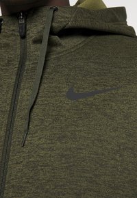 Sweat à capuche zippé vert avec un tissu texturé, présentant un logo Nike noir sur la poitrine et des cordons ajustables sur la capuche.