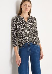 Blusa a maniche lunghe nera con motivo astratto color crema, scollo con bottoni e tessuto morbido, abbinata a jeans larghi blu.