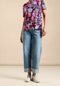 Person som bär en kortärmad topp med rosa blommor, ljusblå uppvikta jeans, vita sneakers och ett armband på vänster handled mot en beige bakgrund.