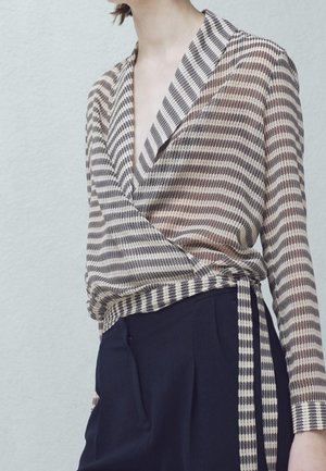 Kobieta ubrana w bluzkę kopertową z długim rękawem w brązowo-biały wzór w kratę, zestawioną z wysokim stanem granatowych spodni.
