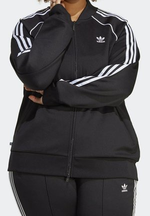 Persoon draagt een zwart Adidas trainingspak met witte strepen, armen gekruist, toont bovenlichaam en middel, op een effen lichte achtergrond.