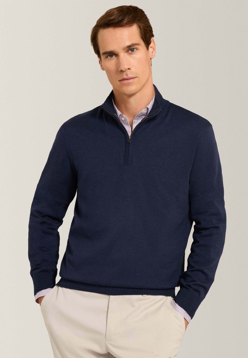 Pull en tricot bleu marine avec un col zippé mi-hauteur, des poignets et un ourlet côtelés, porté par-dessus une chemise à rayures légères. Texture douce, coupe décontractée.