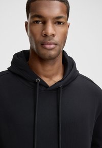 Felpa nera con cappuccio e coulisse, caratterizzata da una texture morbida e un design semplice. Il tessuto è liscio, senza motivi o loghi visibili.