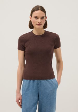 Frau mit kurzen braunen Haaren, die goldene Creolen, ein eng anliegendes dunkelbraunes kurzärmeliges Oberteil und hellblaue Jeans mit hoher Taille trägt.