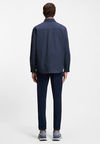 Tamsiai mėlyna marškiniai su ilgomis rankovėmis, nugarinės dalies įsiuvais ir sagomis rankogaliuose, derinami su tamsiai mėlynomis kelnėmis ir mėlynomis sportinėmis bateliais, demonstruojantys aptakų dizainą.