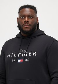 Fekete kapucnis pulóver, puha anyagból készült, elöl kenguruzsebb található, húzózsinórral ellátott kapucnival, és fehér "Tommy HILFIGER" logóval, piros és kék részletekkel.