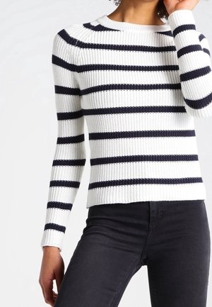 Pull en maille blanc avec des rayures horizontales noires, porté avec un pantalon noir sur une personne, torse moyen visible, un bras levé près du visage.
