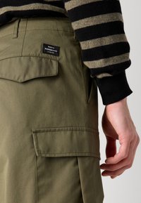Khakifarbene Cargo-Hosen mit seitlichem Taschen und Mads Nørgaard-Label, kombiniert mit einem schwarz-oliv gestreiften Pullover. Glatte Stoffstruktur.
