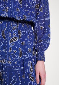 Vestido azul con estampado de paisley, cintura elástica, mangas largas abullonadas y puños fruncidos. Presenta acentos en blanco y negro en todo el diseño.