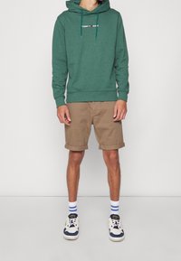 Grön hoodie med en dragkedja på huvan och vit logotyp, kombinerad med bruna korta shorts. Skorna inkluderar vita och marinblå sneakers med accentfärger.