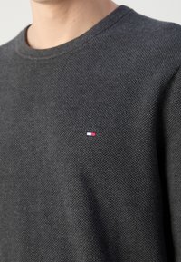 Gros plan d'un pull texturé gris foncé avec un petit logo rectangulaire rouge, blanc et bleu sur la poitrine, porté par une personne.
