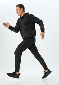 Musta urheiluhuppari, jossa on rento mitoitus, yhdistettynä mustiin kapeisiin joggereihin. Asusteessa on sileät pinnat ja heijastavat yksityiskohdat.