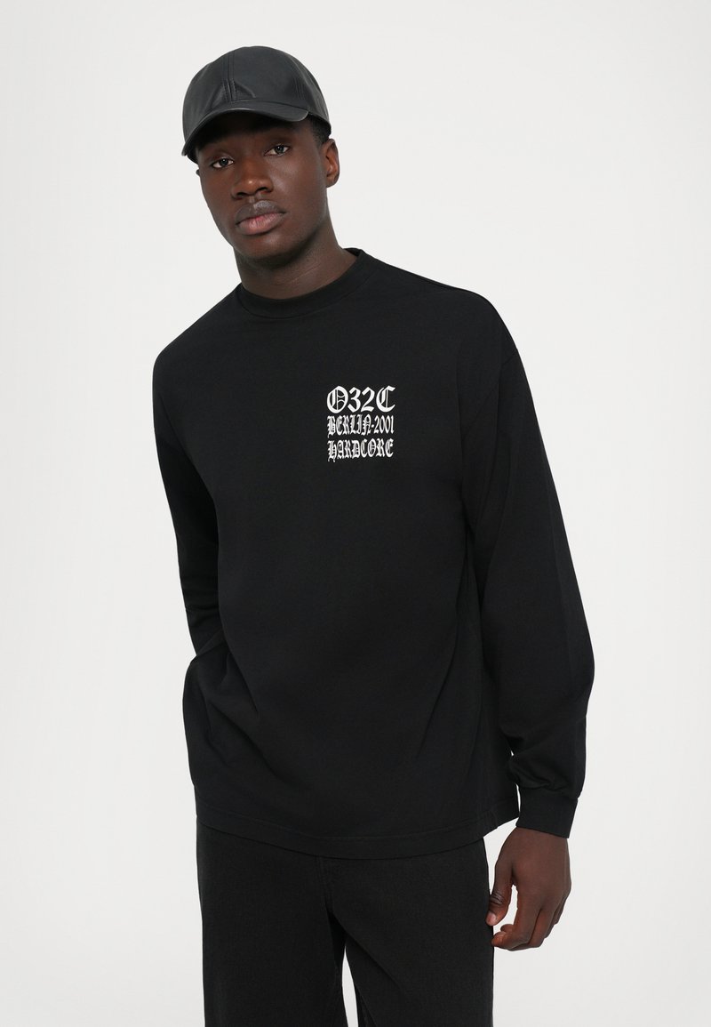 Homme portant une casquette en cuir noir et une chemise à manches longues noir avec un texte gothique blanc indiquant "032C Berlin 2001 Hardcore."