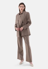 Brauner, maßgeschneiderter Anzug mit einem doppelt geknöpften Blaser und passenden Hosen, aus glattem Stoff gefertigt. Akzentuiert mit eleganten, spitzen Absätzen.