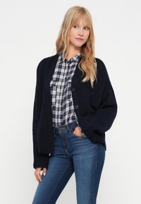 Granatowy sweter zapinany na guziki z długimi rękawami, zestawiony z koszulą w kratę i niebieskimi dżinsami, wyeksponowany na jednolitym tle.