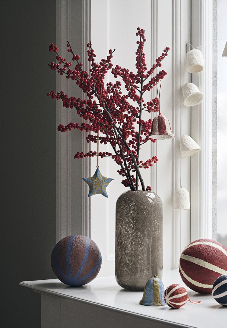 Vase mit roten Beerenzweigen, grau texturierte Oberfläche. Bunte handgemalte Kugeln und Filzornamente hängen in der Nähe eines Fensters.
