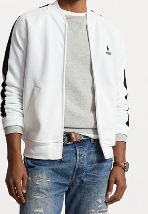 Mann trägt weiße Jacke mit Reißverschluss und schwarzen Streifen an den Ärmeln, graues Sweatshirt, ausgewaschene blaue Jeans, braunen Ledergürtel und Uhr am linken Handgelenk.