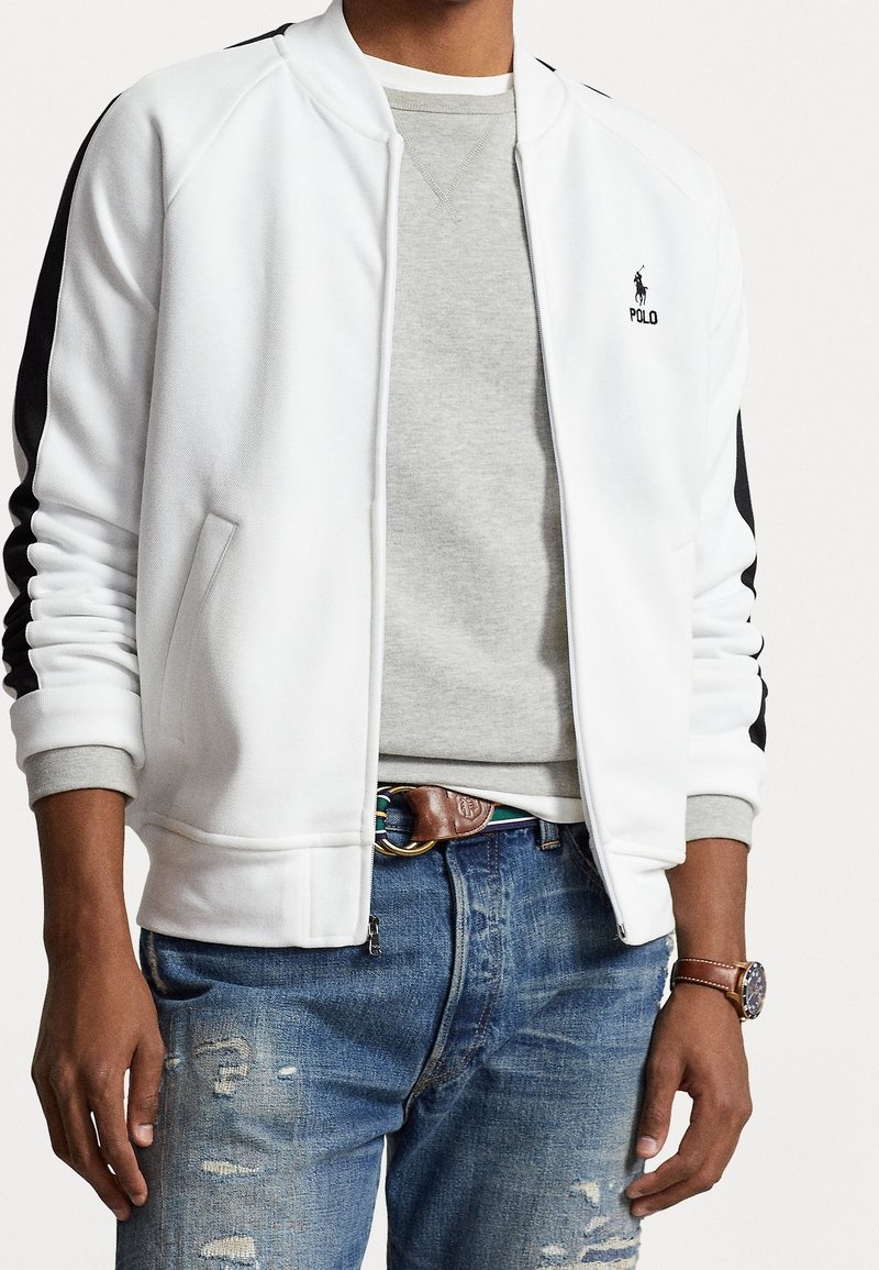 Mężczyzna w białej kurtce na zamek z czarnymi paskami na rękawach, szarej bluzie, zniszczonych niebieskich dżinsach, brązowym skórzanym pasku i zegarku na lewym nadgarstku.