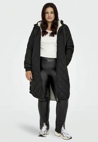 Γυναίκα που στέκεται ντυμένη με ένα μαύρο τζάκετ με κουκούλα, λευκό τοπ, μαύρο παντελόνι δερματίνης και λευκά αθλητικά παπούτσια, σε απλό άσπρο φόντο.