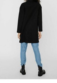 Abrigo largo negro con cuello y ajuste ligeramente holgado, combinado con pantalones azul claro y botas negras hasta el tobillo. Textura suave, diseño minimalista.