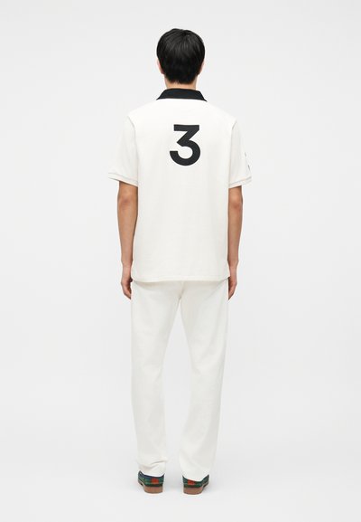 Άνδρας με την πλάτη γυρισμένη, φοράει λευκό πόλο μπλουζάκι με μαύρο γιακά και μεγάλο μαύρο αριθμό 3 στην πλάτη, λευκό παντελόνι και σκουρόχρωμα αθλητικά παπούτσια.