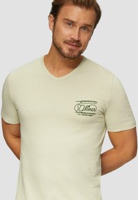 Lichtgroene T-shirt met korte mouwen en V-hals, voorzien van een borstlogo in donkergroen met tekst en een ontwerp van een vistok. Zachte, gladde stof.