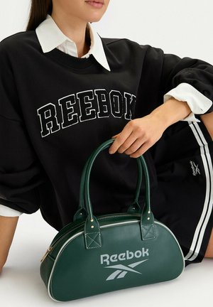 Osoba ubrana w czarną bluzę Reebok i spodenki, trzymająca ciemnozieloną torebkę Reebok z białym logo i złotym suwakiem.