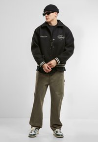 Jachetă varsity din lână neagră cu manșete albe cu dungi și logo brodat; asortată cu pantaloni cargo oliv și adidași verzi cu alb.