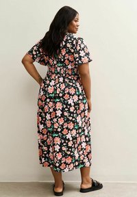 Mujer vestida con un vestido midi negro con estampado floral en tonos rosa y blanco, de pie con una mano en la cadera y usando sandalias negras de tipo deslizante.