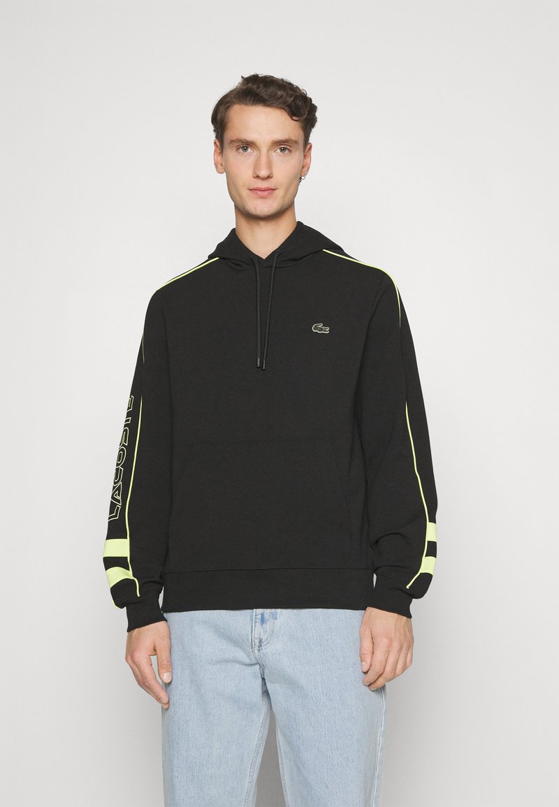 Hoodie noir avec cordon de serrage, présentant des accents jaune néon le long des épaules et des manches. Logo Lacoste sur la poitrine. Tissu texturé.