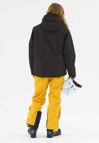 Schwarze Skijacke mit Kapuze, kombiniert mit gelben wasserdichten Hosen. Zubehör umfasst einen weißen Helm, der in einer Hand gehalten wird.