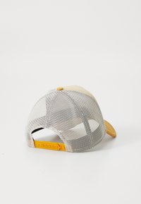Gorra de camionero con un panel frontal beige, acentos amarillos y una parte trasera de malla. Cuenta con una correa ajustable y un detalle en relieve en la parte frontal.