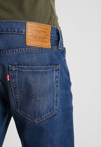 Jeans in denim di colore blu scuro con una toppa in pelle marrone con logo a rilievo. La tasca posteriore ha cuciture a contrasto e un dettaglio a linguetta rossa.