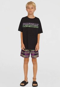 Lyshåret barn står iført en sort, oversize T-shirt med logo, lilla og grønne mønstrede shorts, sorte slidesandaler og ankelarmbånd.