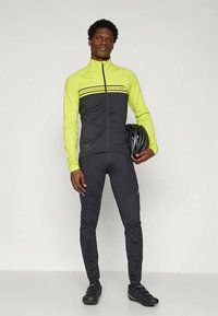 Man wearing black and neon yellow cycling jacket and tights, holding a black bike helmet, standing against a plain background.