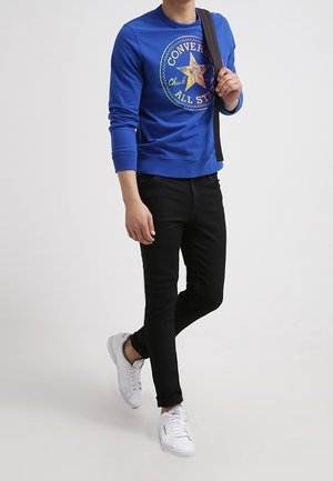 Modrá mikina s grafikou "Converse Chuck All Star"; černé slim-fit kalhoty; bílé tenisky se šňůrkami; neformální outfit.